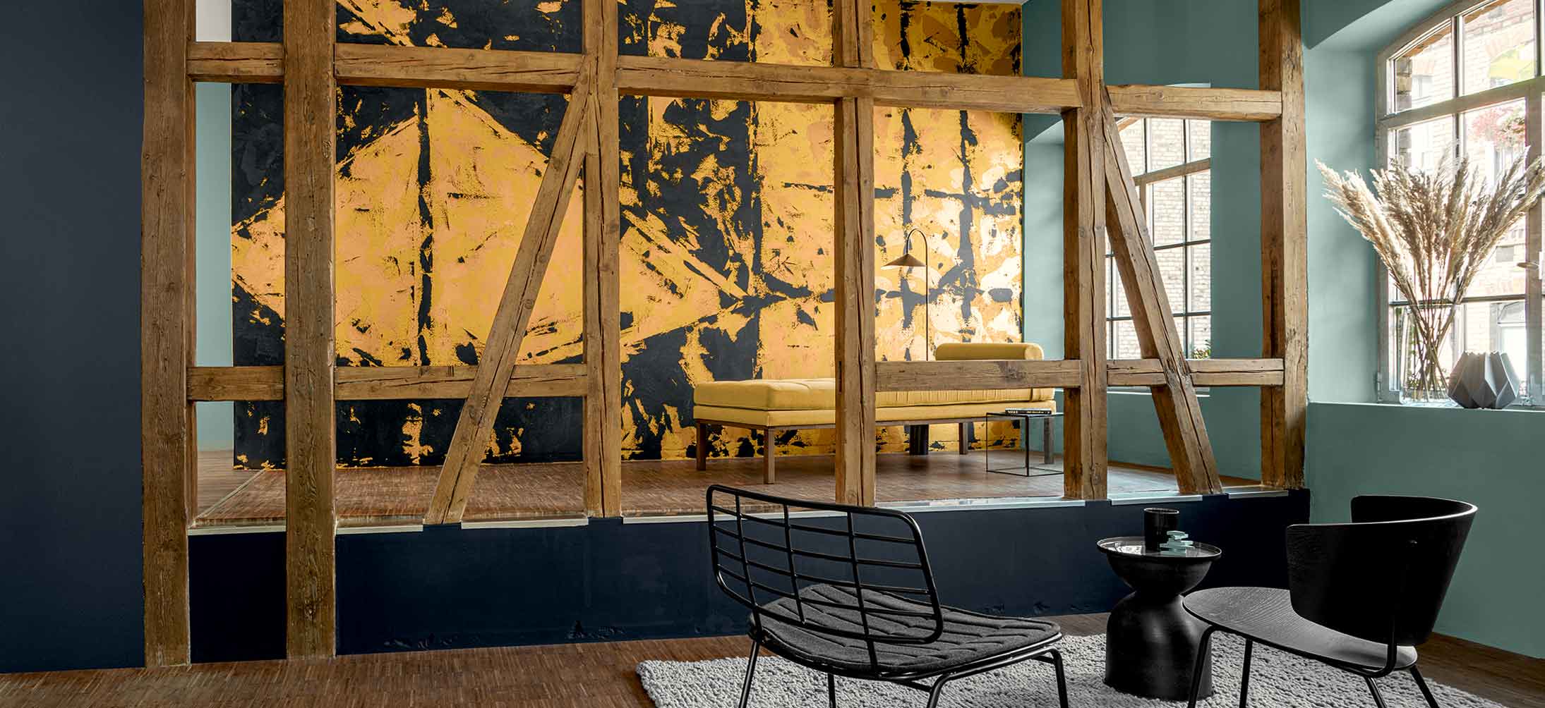 Rustikale Holzbalken im Vordergrund die ein Podest eomrajmem. dahiner eine große in farbigem Design gestrichene Wand in goldgelb/schwarz gehalten