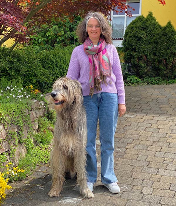 Bernadette Kempf steht mit einem großen, zotteligen Hund am Rande eines Gartens, sie trägt Jeans, lila Pullover und einen Schal